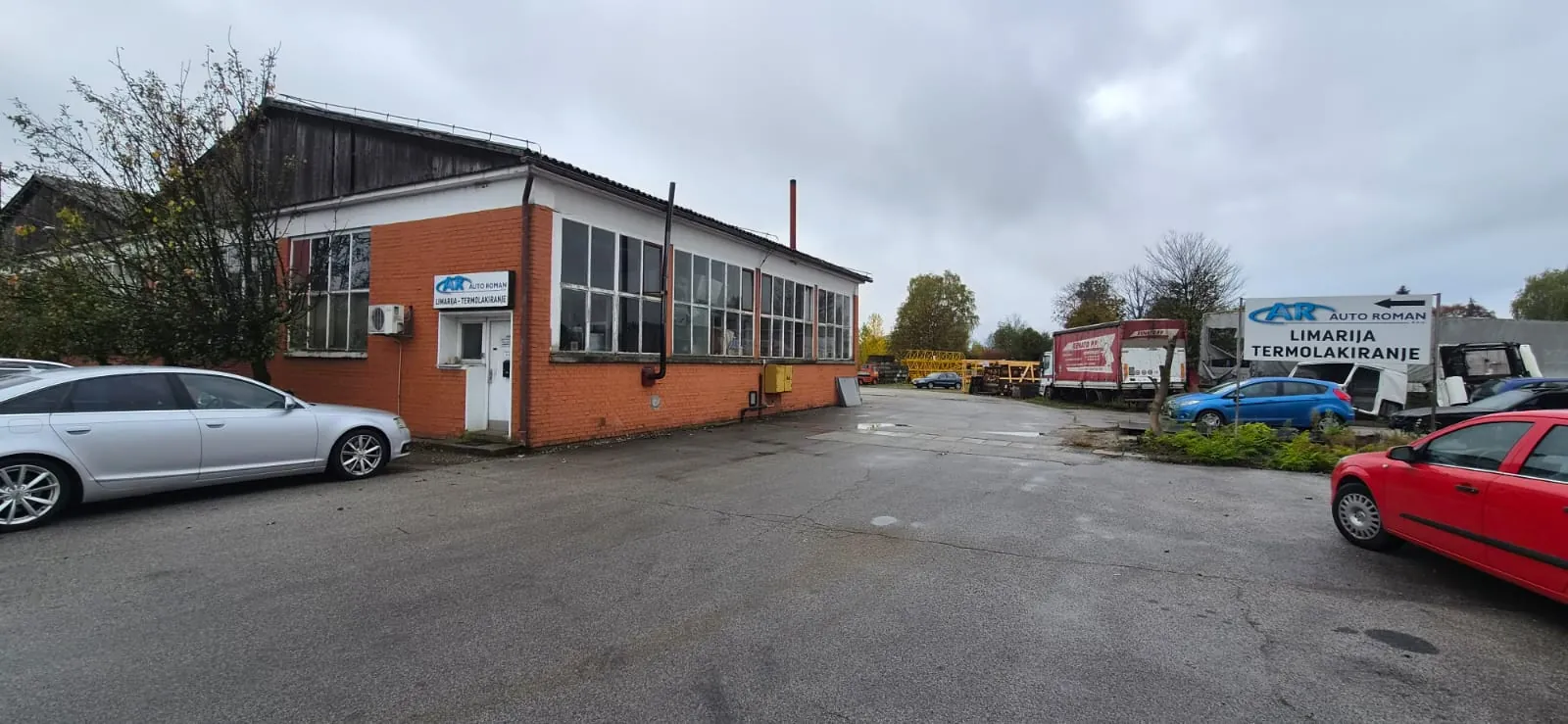 Narančasta zgrada od cigle s natpisom Auto Roman, limarija i termolakiranje, i tabla sa strelicom u dvorištu